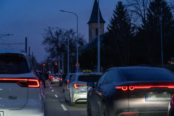 Bratislava city traffic at dusk with cars queuing at red light, Toyota Corolla taxi, Mazda CX-60e and church steeple silhouetted against blue evening sky, tram wires overhead, February 2026