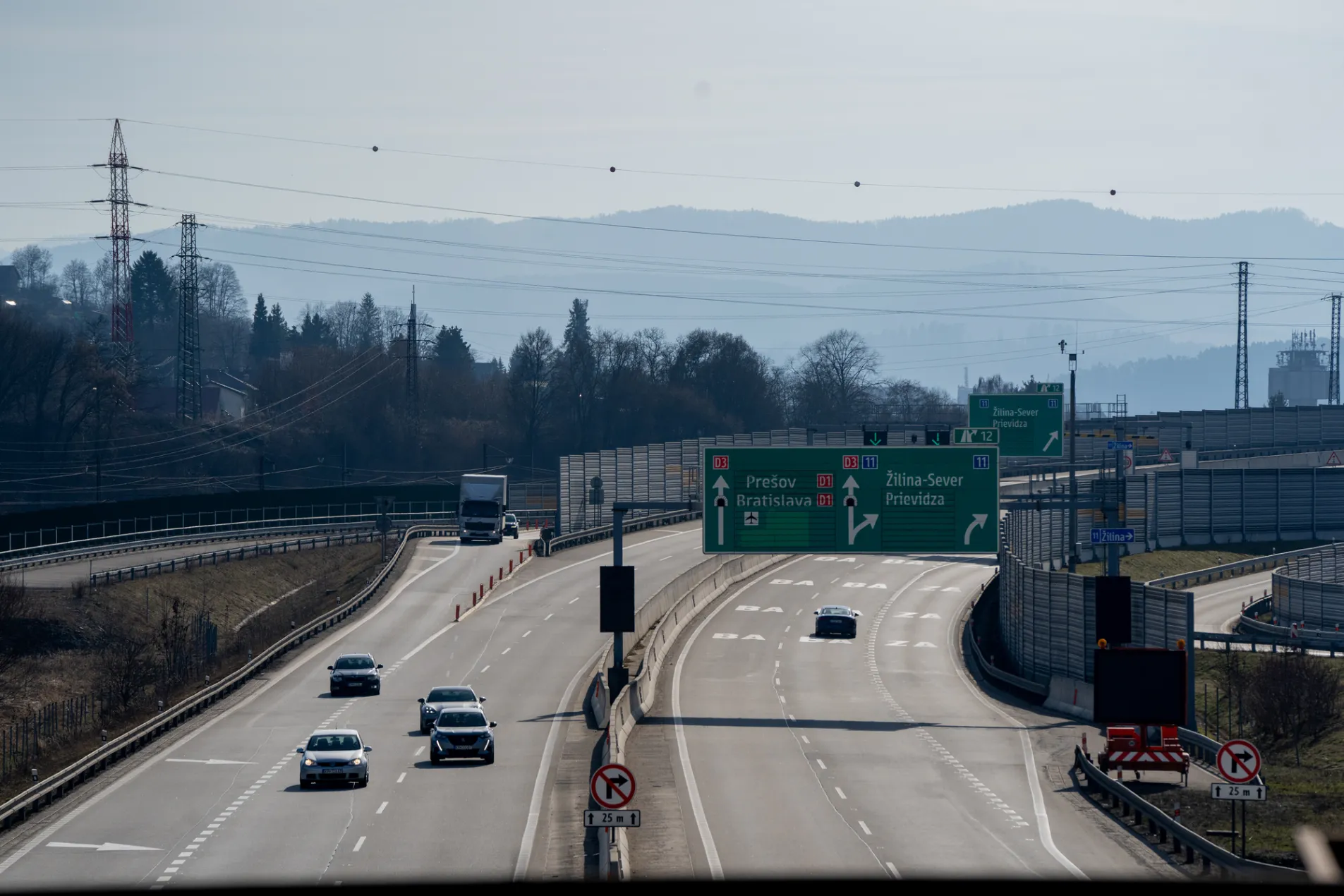 Slovakia D1 D3 motorway junction with green overhead directional signs showing Prešov, Bratislava, Žilina-Sever and Prievidza, multiple lanes with cars and truck, mountain silhouette and noise barriers in background, February 2026