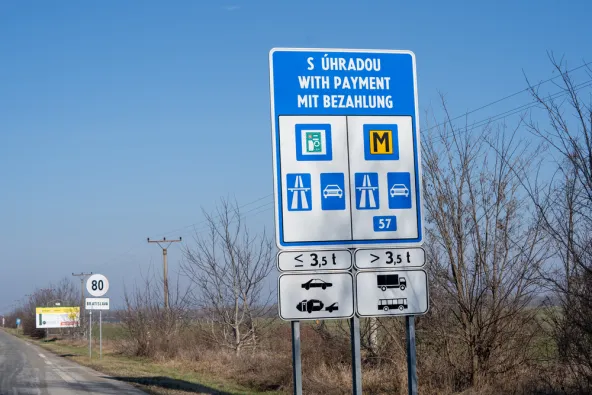 Slovakia blue roadside sign reading S Úhradou / With Payment / Mit Bezahlung showing e-vignette and M motorway toll symbols for vehicles under and over 3.5 tonnes, Bratislava direction sign and 80 km/h speed limit visible in background, February 2026